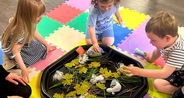 three children around a plastic tray with leaves, twigs and feathers for them to touch